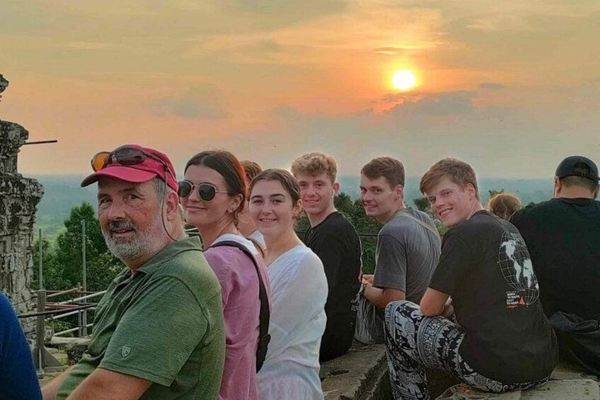 A group of people sitting on a ledge overlooking a landscape with a sunset in the background. 2