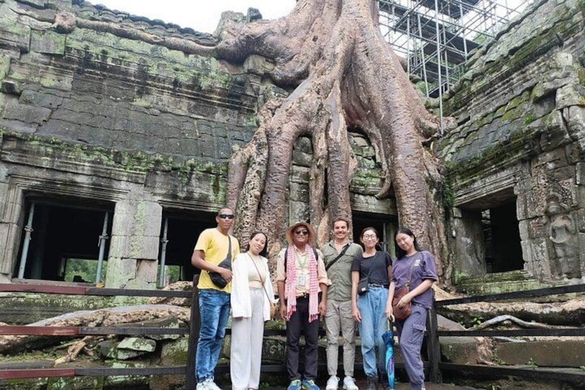 A group of people standing in front of a large tree with roots intertwined with ancient stone architecture. 4