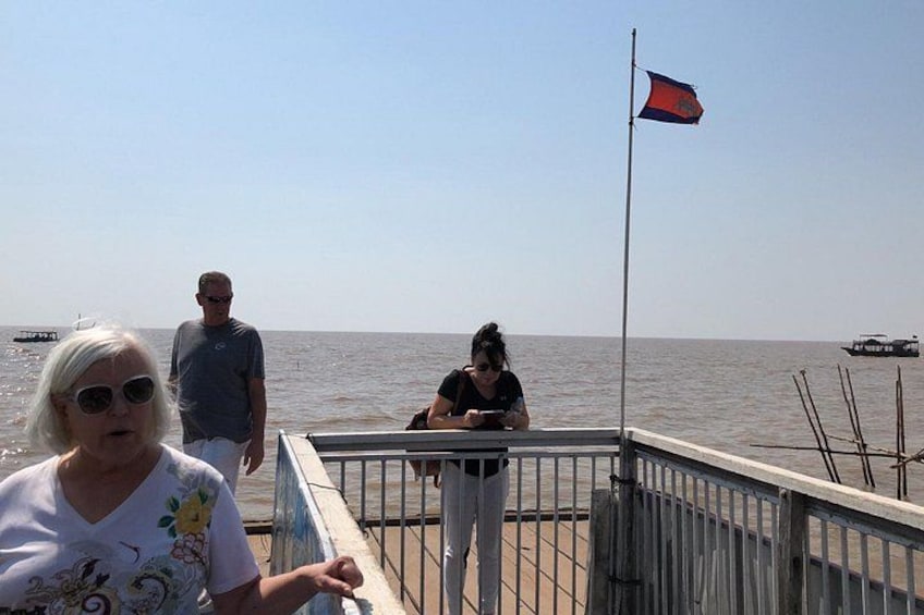 A pier with people standing and walking, a flag on a pole, and a calm sea in the background. 2