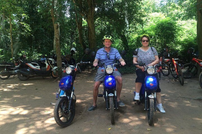 Two people on blue motorcycles in a wooded area with other motorcycles parked around. 1