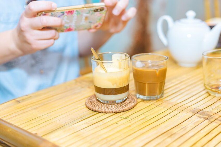 Una persona sosteniendo un teléfono, un vaso con bebidas en capas y una tetera sobre una mesa de madera. 2