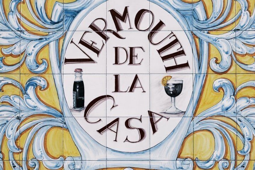 En vägg täckt med kakel, med texten ”VERMOUTH DE LA CASA” och illustrationer av en flaska och ett glas. 1