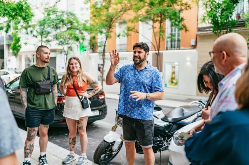 En grupp människor som står på en trottoar, en motorcykel parkerad i närheten och en byggnad i bakgrunden. 2