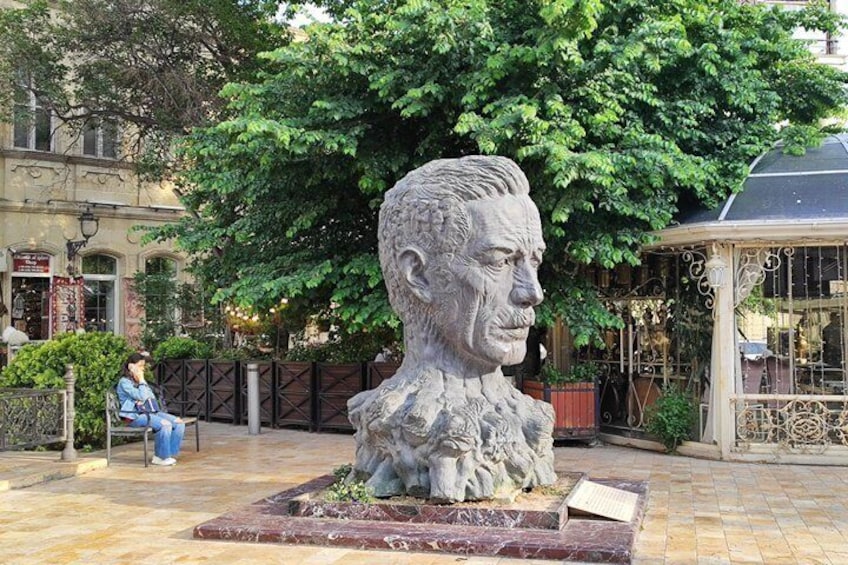 Una escultura de busto en un patio con un árbol, un pabellón y una persona sentada en un banco. 2