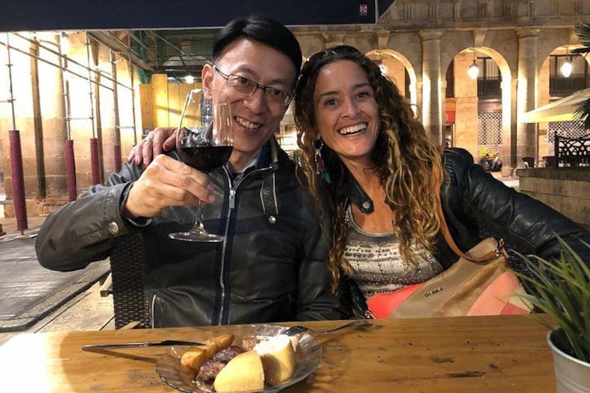 Deux personnes sont assises à une table en bois, devant un verre de vin rouge, un panier de nourriture et une plante en pot. 4