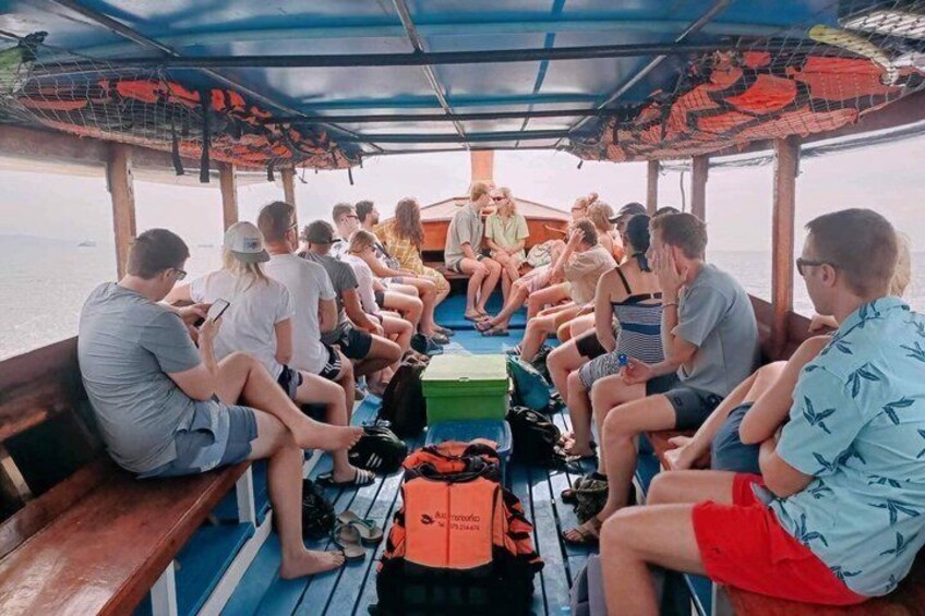 A group of people seated on a boat's deck, some with backpacks, under a covered area with a patterned roof. 3