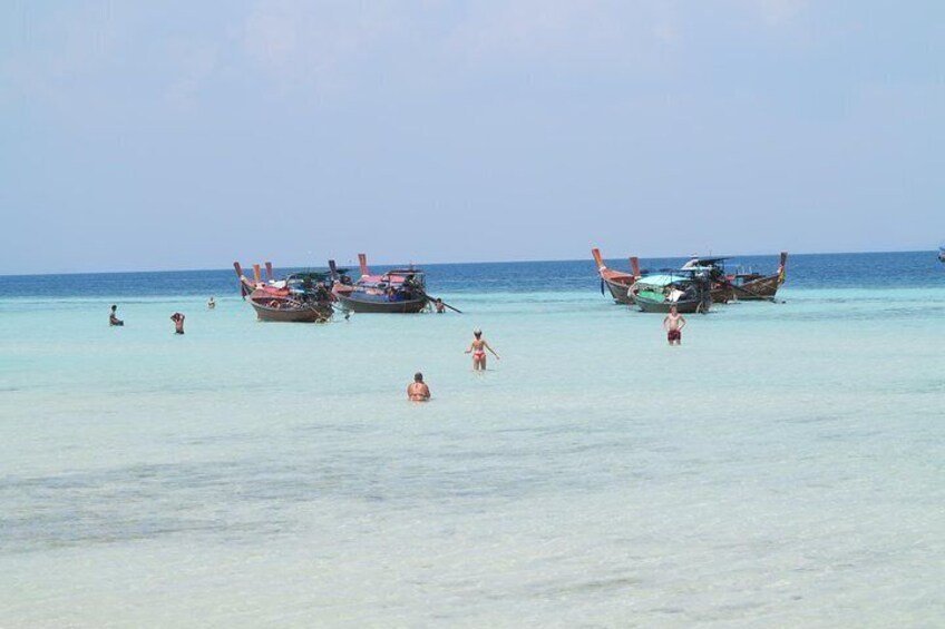 A beach with clear turquoise water, several people swimming and walking, and traditional boats anchored in the distance. 1