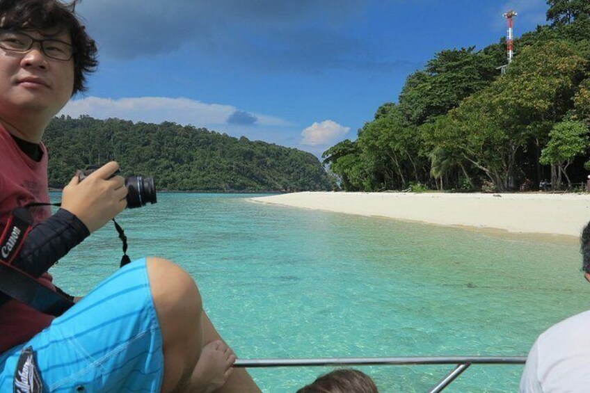 A person taking a photo with a camera, a beach with clear turquoise water, and a lighthouse on a hill. 5