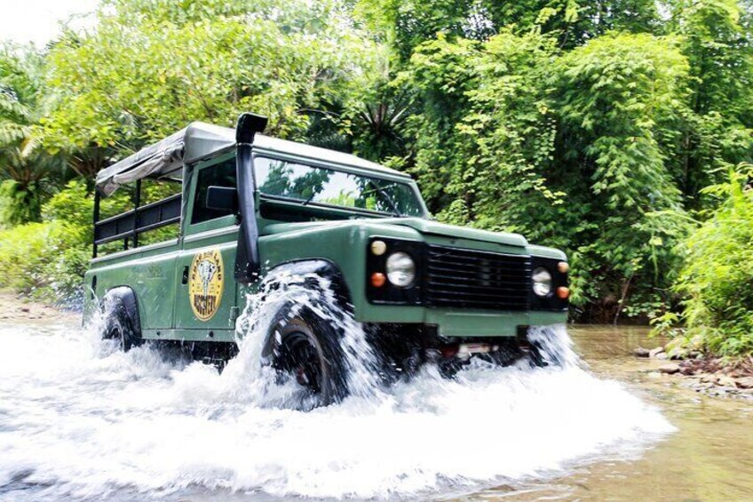 A green off-road vehicle driving through a shallow river surrounded by dense greenery. 1