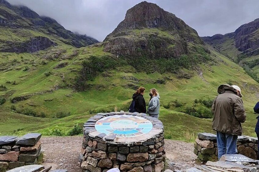 A stone monument with a circular top, surrounded by a stone wall, set in a mountainous landscape with green hills and a prominent rocky peak in the background. 2
