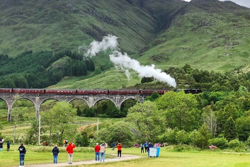 A train crossing a bridge, steam rising, surrounded by lush green hills and a group of people observing. 4