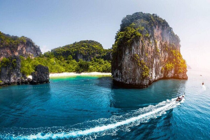 A clear blue sea with a boat creating a wake, surrounded by large limestone cliffs and lush greenery. 2