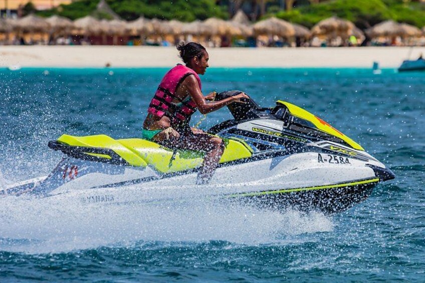 A person riding a yellow and white jet ski on a body of water with a beach and thatched-roof structures in the background. 4