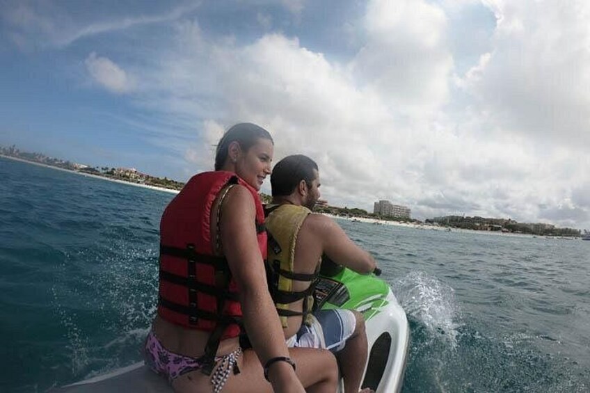 Two people on a jet ski, wearing life jackets, with a coastline and buildings in the background. 3