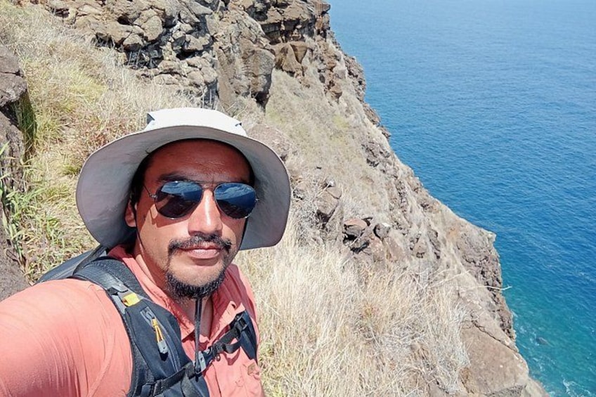 Une personne portant un chapeau et des lunettes de soleil, debout sur une falaise rocheuse, avec une étendue d'eau en arrière-plan. 2