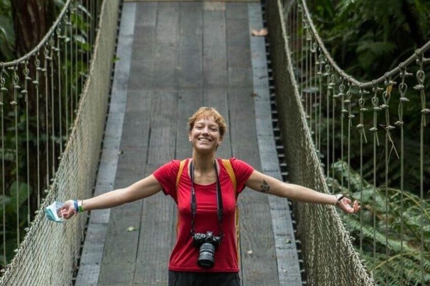 Eine Person steht auf einer Hängebrücke, die Arme ausgebreitet, umgeben von viel Grün. 4