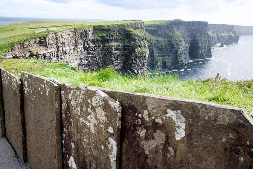 A coastal cliff with a deteriorating concrete barrier, overlooking a body of water and green landscape. 1
