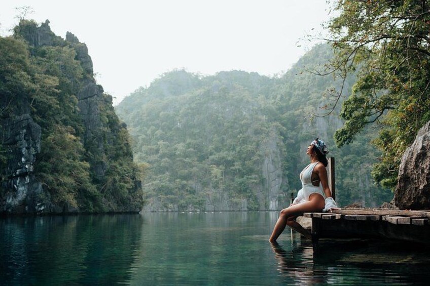 A person sitting on a dock by a calm lake surrounded by forested hills. 2
