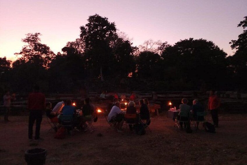 A group of people gathered outdoors at dusk, some seated and some standing, with a fence and trees in the background. 1