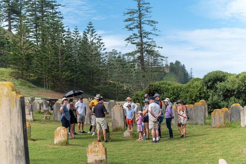 A group of people standing in a cemetery with headstones, surrounded by trees and a grassy hill. 1