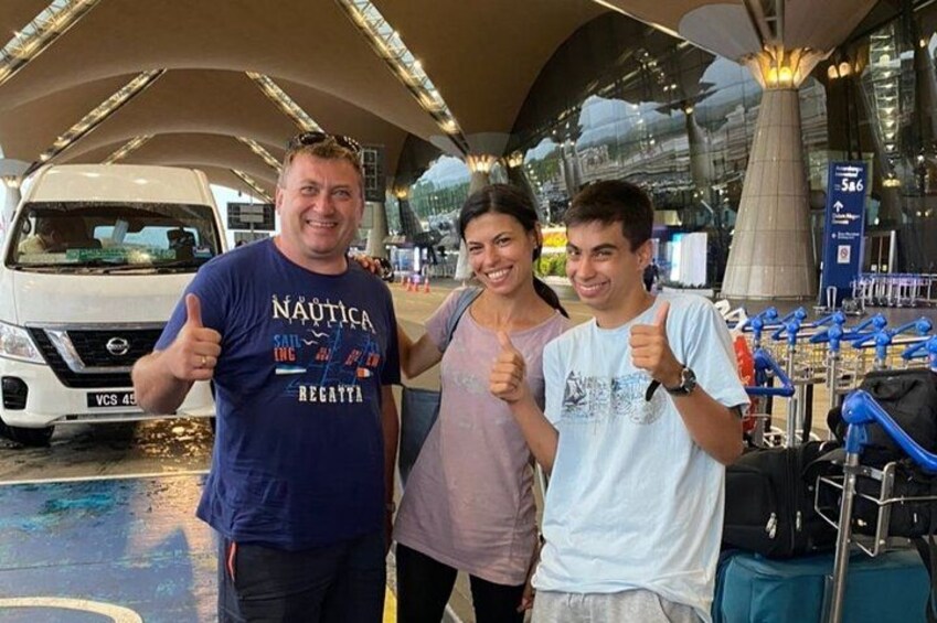 Three people giving thumbs up, a white van, and luggage racks in a terminal. 2