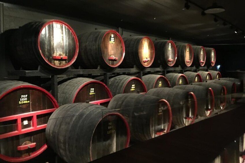 Rows of wooden wine barrels with red rims and labels, arranged in a dimly lit storage room. 1