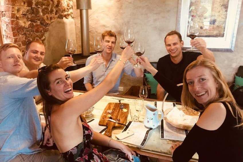 A group of people toasting with wine glasses around a table with a pizza and a cutting board. 5