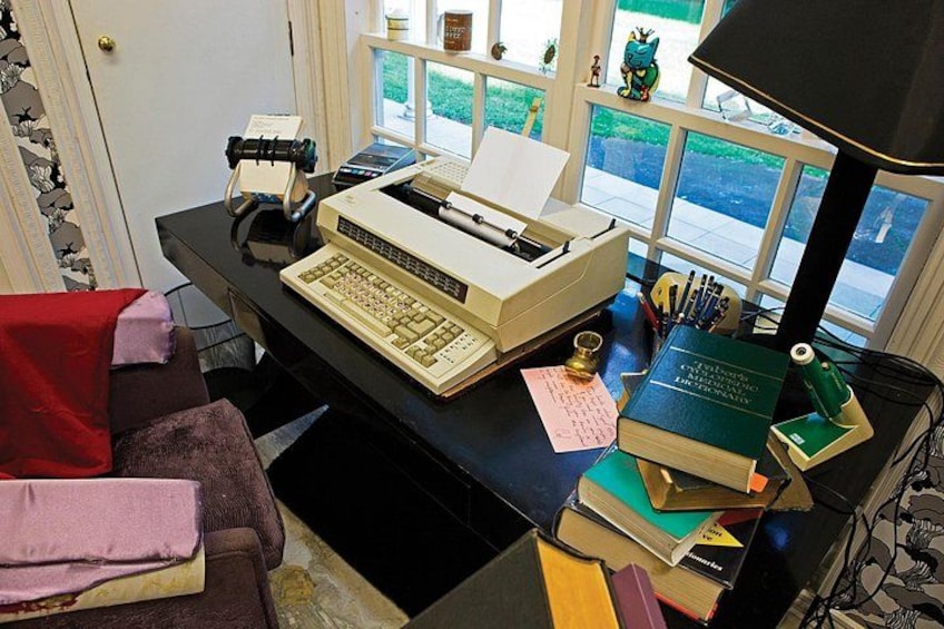 Eine Vintage-Schreibmaschine steht auf einem Schreibtisch mit Büchern, einer Lampe und einem Fenster mit Blick ins Freie. 3