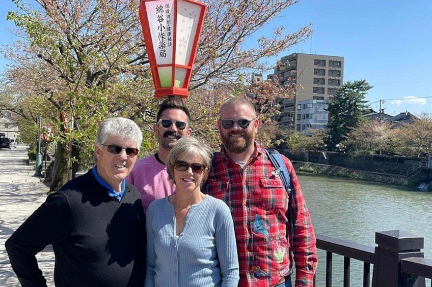 A group of people standing by a river with cherry blossoms and a traditional Japanese lantern in the background. 2