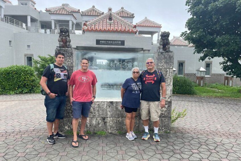 Four people standing in front of a historical monument with a plaque, surrounded by a paved area and buildings with tiled roofs. 1