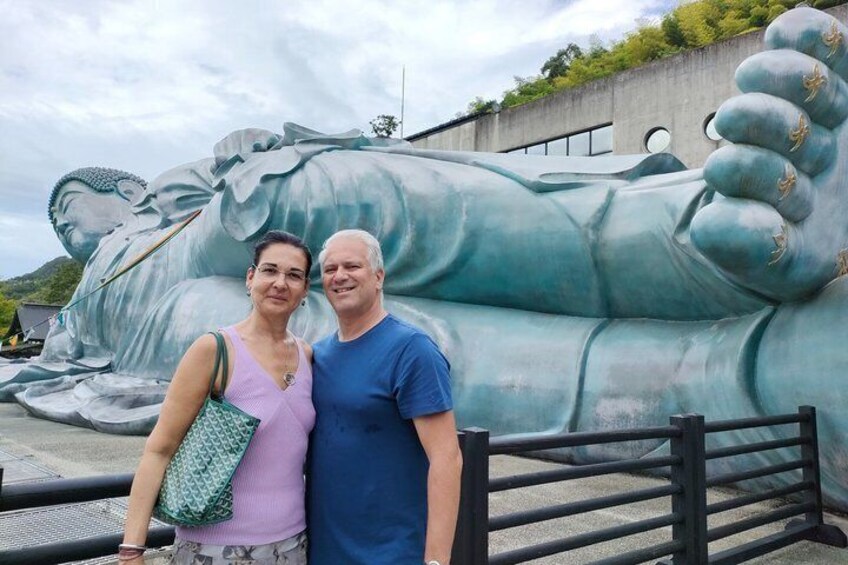 Une grande sculpture représentant une figure allongée, une main levée, une personne debout devant, et une clôture au premier plan. 1