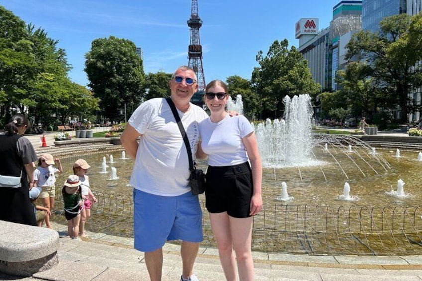 A couple standing in front of a fountain with water jets, a park with trees, and a tower in the background. 1