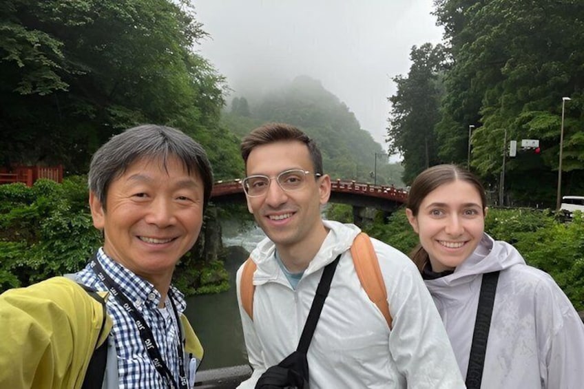 Three people in a forested area with a bridge in the background. 1