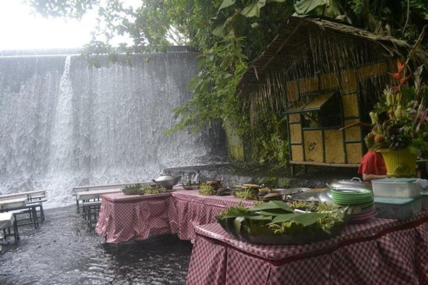 A waterfall with tables set up for dining, surrounded by lush greenery and a thatched-roof structure. 3