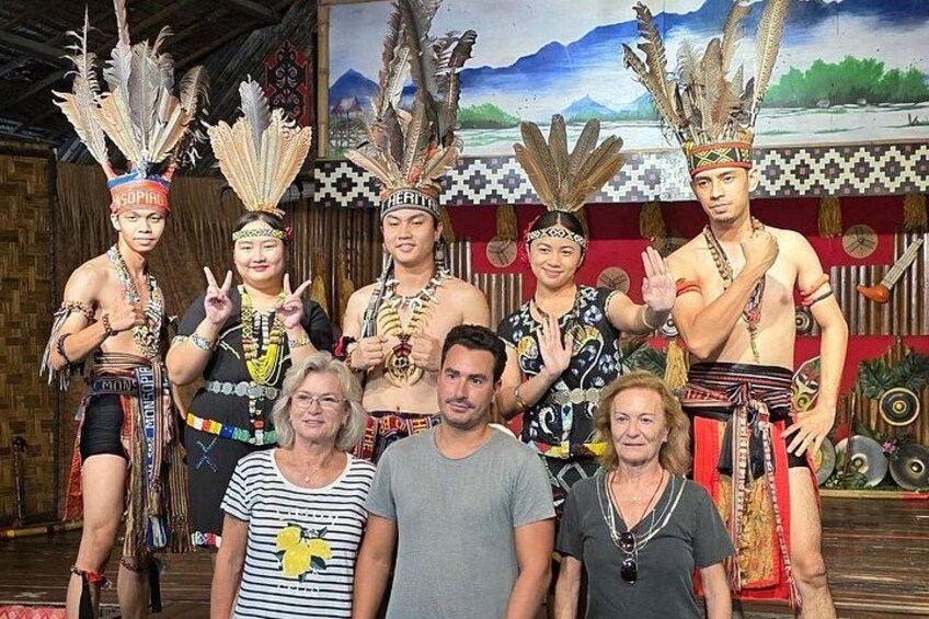 Un gruppo di persone in abiti tradizionali con piume ornate per la testa, in posa di fronte a uno sfondo con un dipinto che raffigura un paesaggio. 1