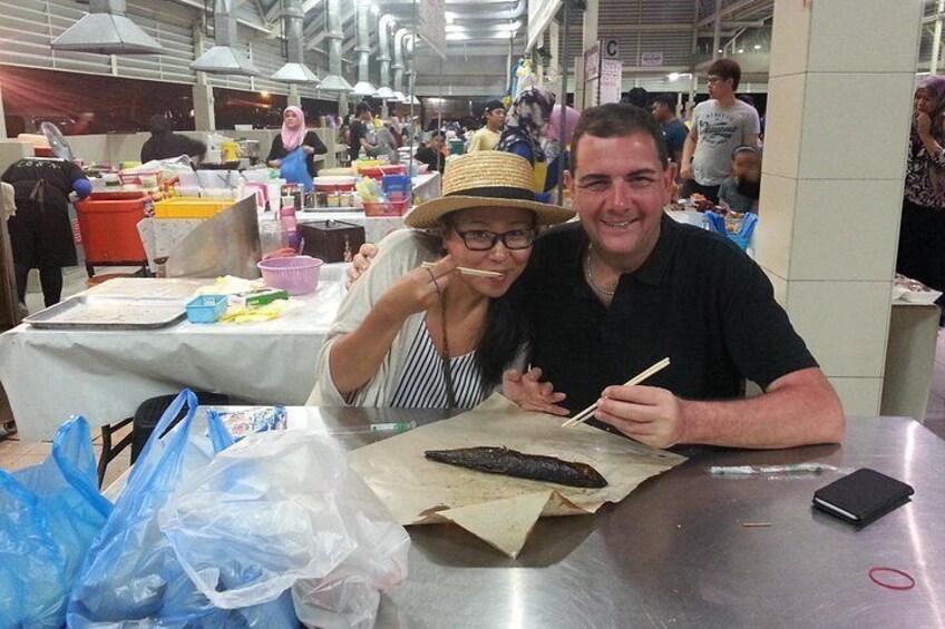 Two people sitting at a table with food items and a fish on it. 2
