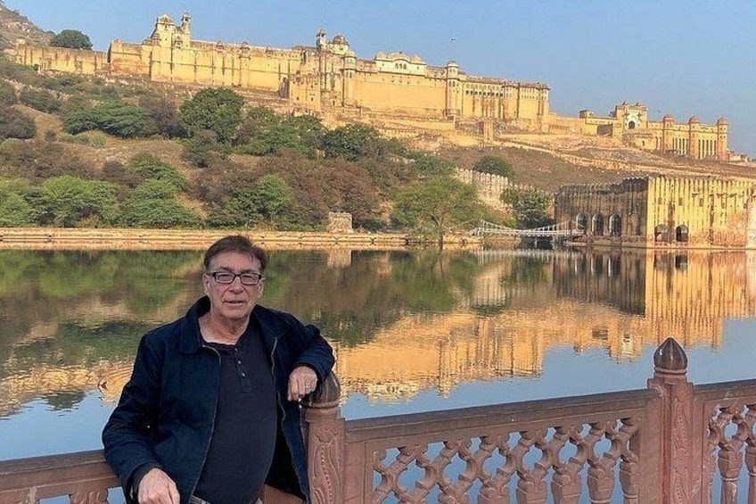 Un homme se tient près d’une étendue d’eau qui reflète la lumière, avec un fort historique en arrière-plan. 1