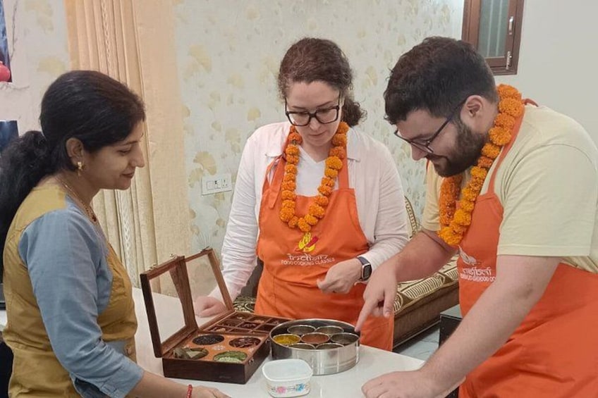 Drei Personen mit orangefarbenen Schürzen und Blumenketten, die mit einer Holzkiste voller Lebensmittel interagieren. 2