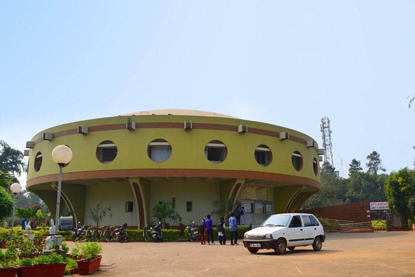 A circular building with a yellow facade and multiple windows, surrounded by parked vehicles and people. 1