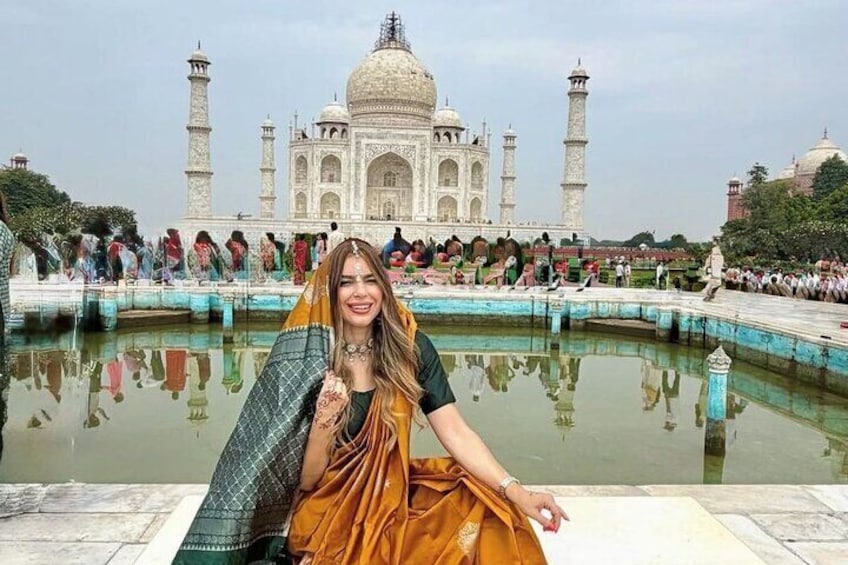 Una persona vestida con atuendo tradicional sentada junto a un estanque que refleja el Taj Mahal al fondo. 1