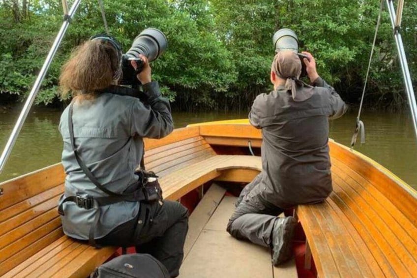 Two people on a wooden boat, each holding a camera, with dense greenery in the background. 3