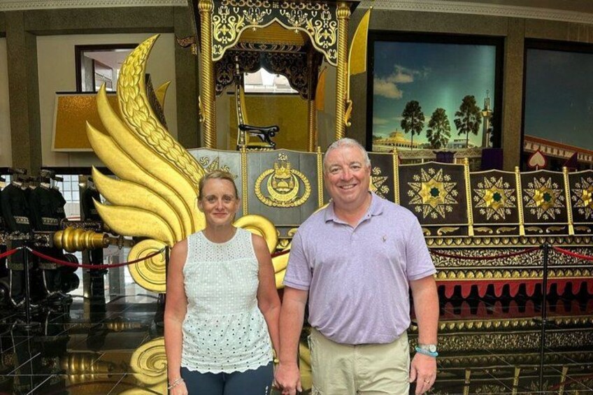 A man and a woman standing in front of a golden throne-like chair with intricate designs. 2