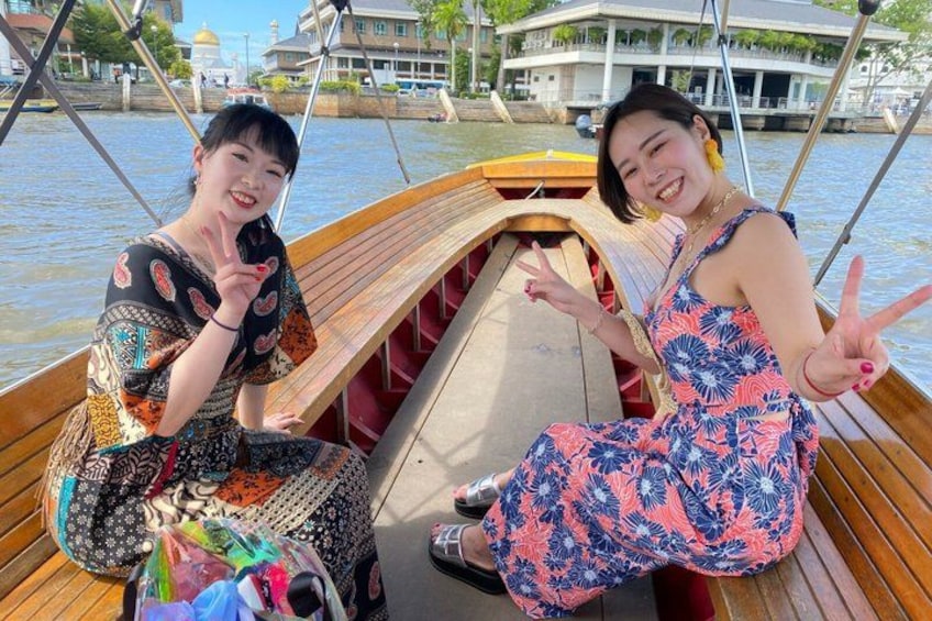 Two people on a wooden boat with a river and buildings in the background. 5