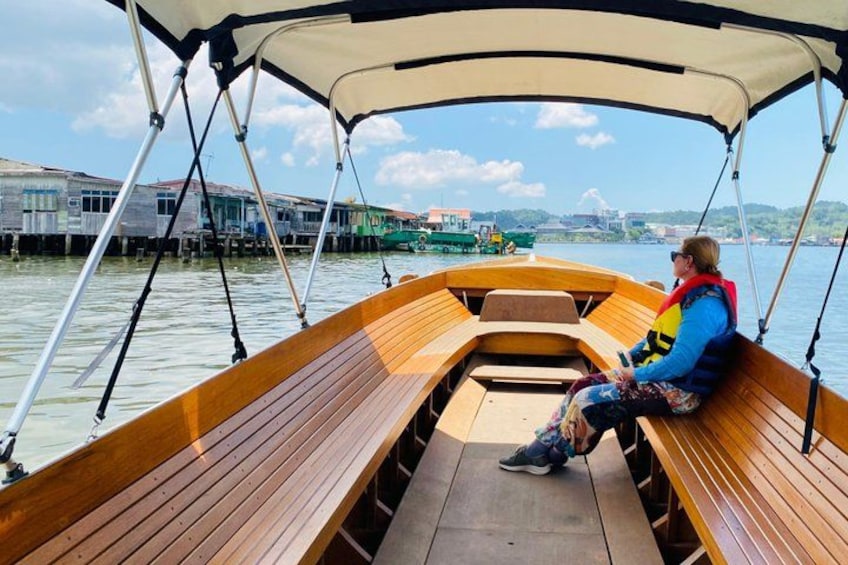 A wooden boat with a canopy, a person sitting on the bench, and buildings along the water. 1
