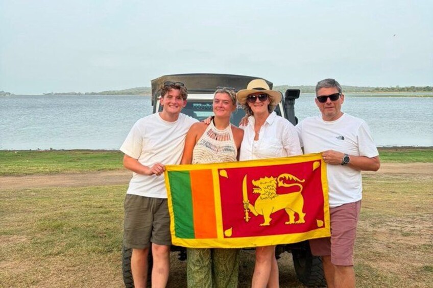 A group of people holding a flag with a lion emblem, standing by a vehicle near a body of water. 4