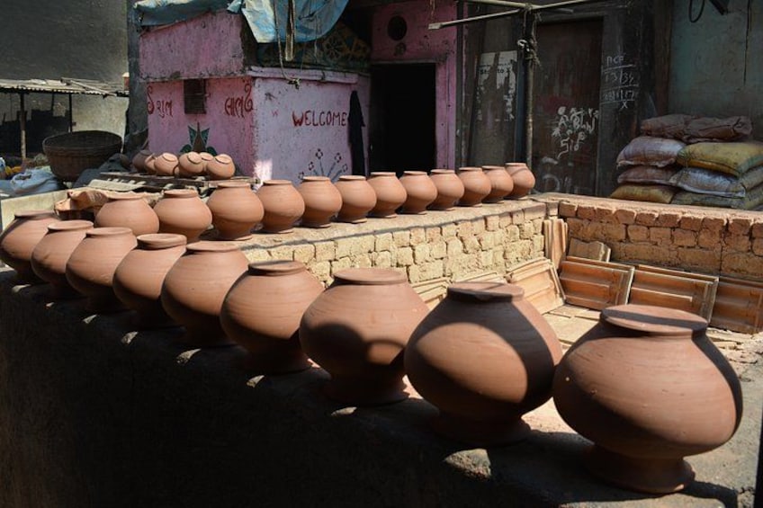 Terracotta pots lined up outdoors, a pink building with 'WELCOME' written on it, and sacks stacked in the background. 2