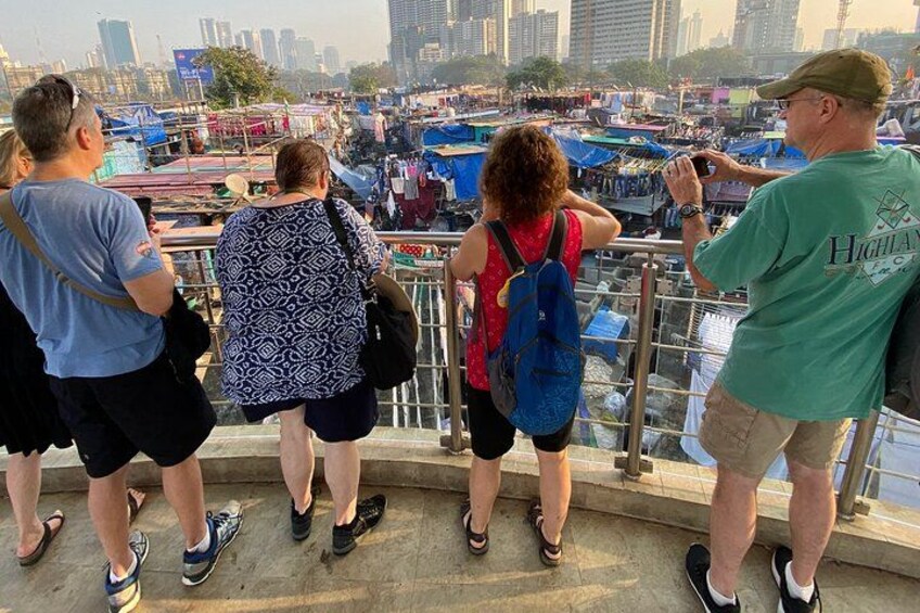 Un grupo de personas de pie en un balcón con vistas a un mercado concurrido, con varios puestos y edificios al fondo. 2