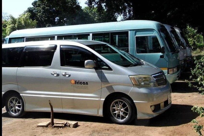 A silver van with 'African' branding parked on a dirt surface, with a blue bus in the background. 1