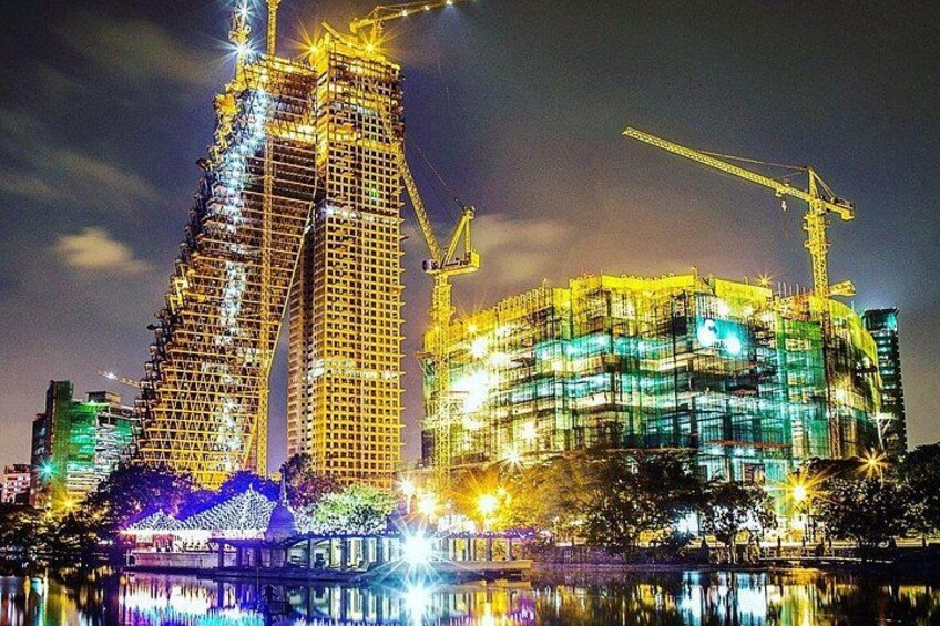 A nighttime cityscape with two illuminated skyscrapers under construction, a bridge, and a body of water reflecting the lights. 5