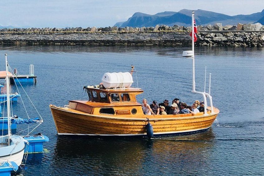 A wooden boat with passengers, docked at a harbor with other boats and a rocky shoreline in the background. 4